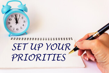 A woman's hand writes the text on a notepad Set your priorities. White background with a clock....