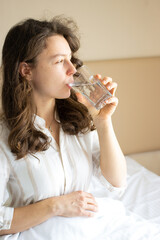 Portrait of young brunette woman drinking pure mineral water, holding transparent glass and looking in distance. Feeling thirsty after waking up. Morning routine, good healthy lifestyle habits.