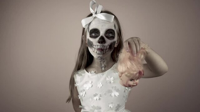 A little girl in a dress with a painted face holds in her hand the bloody head of a doll by her hair