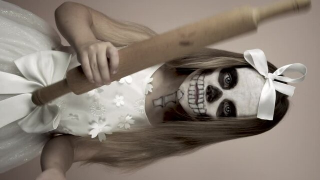 A little girl with a painted face holds a club in her hands. She scares and waves the rolling pin.