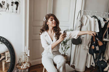 Woman in stylish white outfit takes photos of shoes. Curly dark-haired lady in blouse and pants sits on chair and holds phone in light cozy room