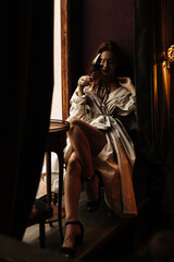 Woman in beige trench sitting in gorgeous bar. Red-haired beautiful lady in white outfit poses near window