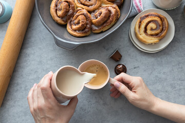 Breakfast scene of a white woman pouring a jar of milk into a cup of coffee with some homemade delicious cinnamon rolls around.