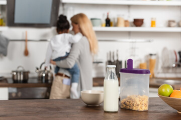 bottle of milk near corn flakes in container, bowl and fruits with blurred mother and adopted african american kid on background