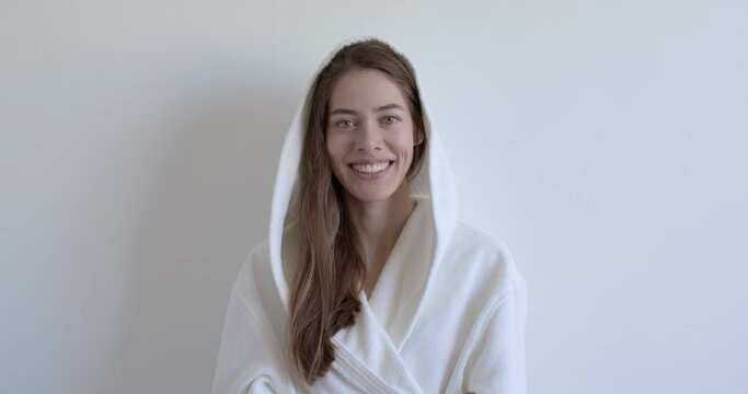 Good-looking female in white bathrobe smiling at camera having perfect toothy smile, long hair, Attractive lady posing on white studio background, in morning at weekends, relaxed lady portrait