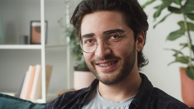 Young brunette man smiling portrait, guy with beard wearing circle glasses, sitting on sofa in the living room. Businessman looking in camera, posing at home, wide smile, teeth with braces close-up. 