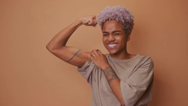 Positive smiling African American young woman raises muscular arm shows biceps has powerful look smiles happily dressed in casual t shot, isolated over brown background, says look how strong I am.