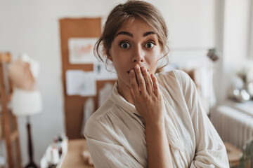 Dark haired lady looks into camera in surprise. Shocked woman in white blouse poses in office and covers her mouth