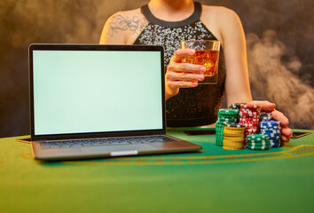 Young woman celebrates winning at an online casino. There is a laptop on the table, a lot of colored chips. A woman holds a glass of alcohol in her hands. Gambling business, risk, passion.
