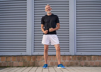 Middle aged athletic man in sportswear holding skipping rope while standing outdoors over grey background