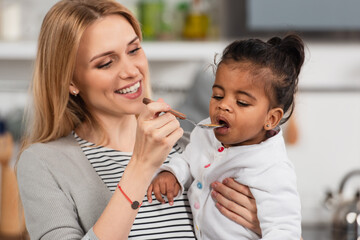 cheerful mother holding spoon and feeding adopted african american daughter
