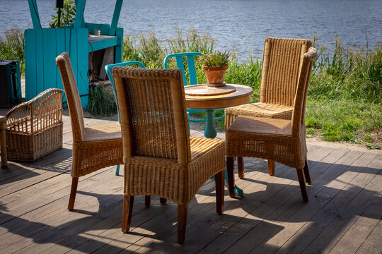 A wooden table and four wicker chairs - garden furniture standing on a bright wooden patio, on the riverbank. Sunny, summer day. 
