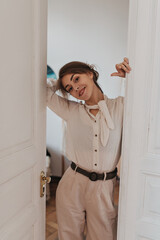 Smiling lady in beige pants and blouse leaning on white door. Happy charming woman in stylish suit sincerely smiles in cozy room