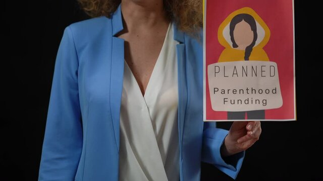 Unrecognizable Female Speaker Holding Planned Parenthood Funding Banner In Hand Talking Standing In Camera Flashes At Black Background. Woman Rising Attention To Social Problem