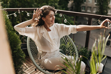 Brunette woman takes selfie on balcony. Portrait of happy cheerful lady in beige blouse and pants...