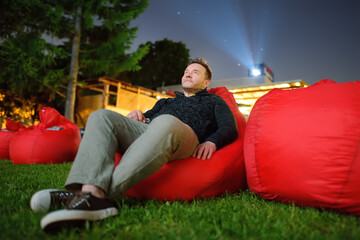 Handsome man laying on pillow on grass and watching movie at outdoor cinema in public park. Perfect...