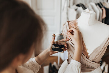 Brunette woman pinning up lace to dress. Dark-haired lady designs beautiful festive beige outfit in office. Blur effect