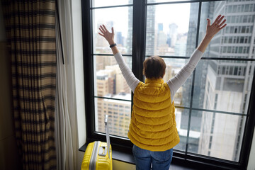 Traveler with suitcase looks and admires of amazing view the skyscrapers of Manhattan outside window. Woman tourist stayed in hotel room in New York. Tourism and travel in USA.