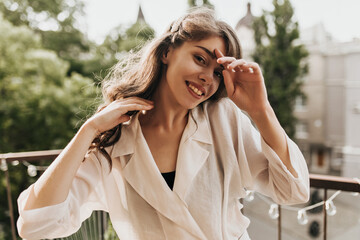 Young lady in beige outfit smiling and looking at camera on terrace. Charming pretty woman in white...