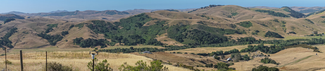 Fototapeta na wymiar Cambria, CA, USA - June 8, 2021: Panorama landscape of hills and farm in Back country with dry ranch land and patches of green trees under blue sky.