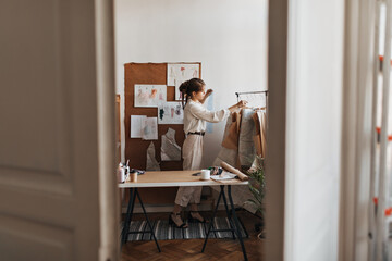 Fashion designer working at her office. Stylish young woman in beige pants and blouse poses in cozy room full of garments patterns and samples