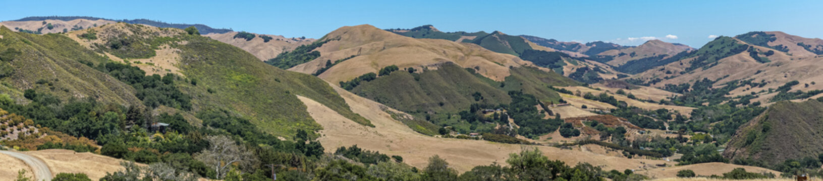Cambria, CA, USA - June 9, 2021: Panorama landscape of Back country with dry ranch land and patches of green trees on steep flanks of mountains under blue sky.