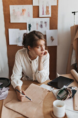 Young woman paint draws sketch. Dark-haired lady in beige stylish blouse thoughtfully poses in cozy office