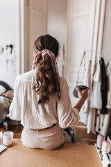 Woman with ponytail sitting on table and holding coffee. Charming lady wth wavy hair drinking tea...