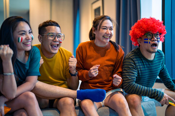 Group of Asian people friends sit on sofa watching and cheering sports games competition on TV together at home. Excited man and woman sport fans celebrate sport team victory in national sports match