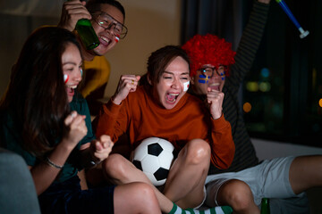 Group of Asian people friends sit on sofa watching and cheering sports games competition on TV together at home. Excited man and woman sport fans celebrate sport team victory in national sports match