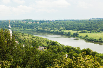 Obraz premium A church hidden by trees on a hill by the river. On both sides of the river there is a green forest.
