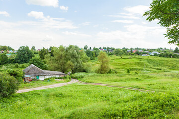 Rustic house with a gray roof against the background of green fields, grass and trees. Russian village.