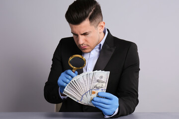 Expert authenticating 100 dollar banknotes with magnifying glass at table on light grey background. Fake money concept