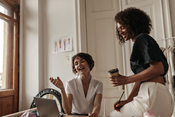Fototapeta premium Pretty short-haired women in stylish blouses talk, smile and look at laptop screen in office. Curly dark-skinned woman in black top holds coffee cup.