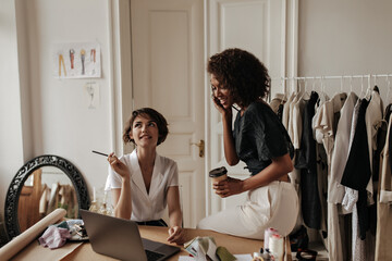 Creative young fashion designers creating new clothes in cozy office. Short-haired Caucasian woman thoughtfully looks up and holds pencil. Dark-skinned lady holds coffee cup.