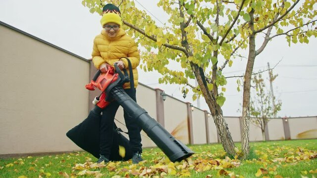 Smiling boy in reading glasses and yellow jacket helping at backyard on cloudy fall day. Slow motion childhood 4K. Teenage boy vacuuming yellow dry leaves from green lawn, having fun times