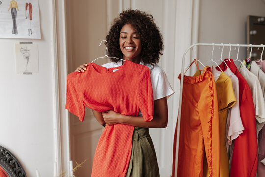 Excited young brunette curly dark-skinned woman in khaki shorts and white blouse smiles sincerely, holds hanger with red dress and poses in dressing room.