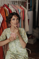 Tanned young attractive woman in green floral dress looks up, holds phone in pink case and sits on chair in cozy dressing room.