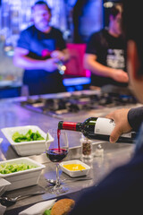 Hand serving a drinking glass of red wine with a bottle in a large metal table in a kitchen with more people and blue light