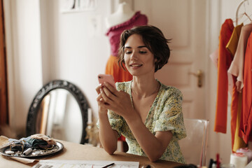 Cheerful tanned brunette short-haired woman in floral v-neck dress chatting on phone, sits at desk...