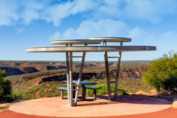 Shade shelter with benches set in the sandstone cliffs of the Kalbarri National Park surrounded by native flora under a blue sky with clouds in Western Australia.
