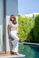 Asian woman is closing her eye and lean against the wall beside the pool and tree background for portrait camera shooting.
