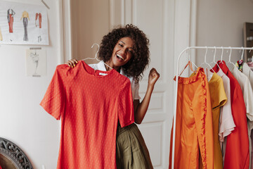 Joyful energized brunette curly dark-skinned woman in khaki shorts rejoices and holds hanger with red dress in dressing room. - obrazy, fototapety, plakaty