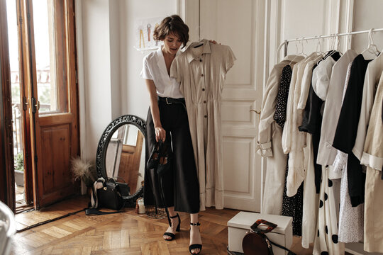 Stylish attractive short-haired woman in midi black skirt and white blouse poses in dressing room, holds trendy linen beige dress and shoes.