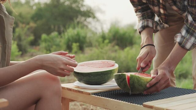 man is cutting a watermelon for in a camping. girlfriend encouraging her boyfriend to prepare fruit. concept of relaxation and recreation.