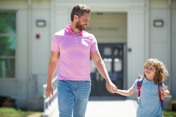 Obraz premium Parent and pupil of primary school go hand in hand. Teacher in t-shirt and cute schoolboy with backpack near school park.