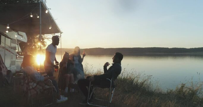 Summertime, group of young people are enjoying a party in nature near the lake, people communicate and relaxing in nature near the camping car, sunset, view from height.