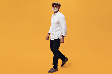 Charming man in white shirt and black pants walking on orange background. Stylish adult with beard and tattoo posing on camera in fashionable clothes