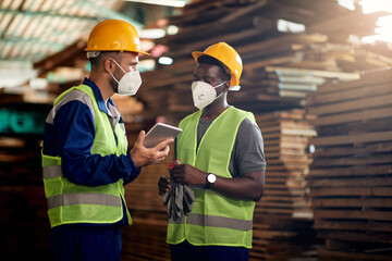 Warehouse foreman and worker with face masks using touchpad and talking while working at lumber warehouse.