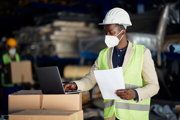 African American warehouse manager works on laptop and wears face mask due to coronavirus pandemic.